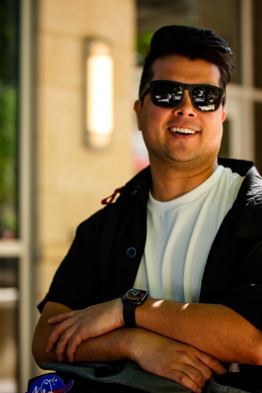 A man with dark hair wearing sunglasses, a white shirt, and a black jacket stands outdoors, smiling with his arms crossed. He is also wearing a smartwatch and is in front of a blurred building background.