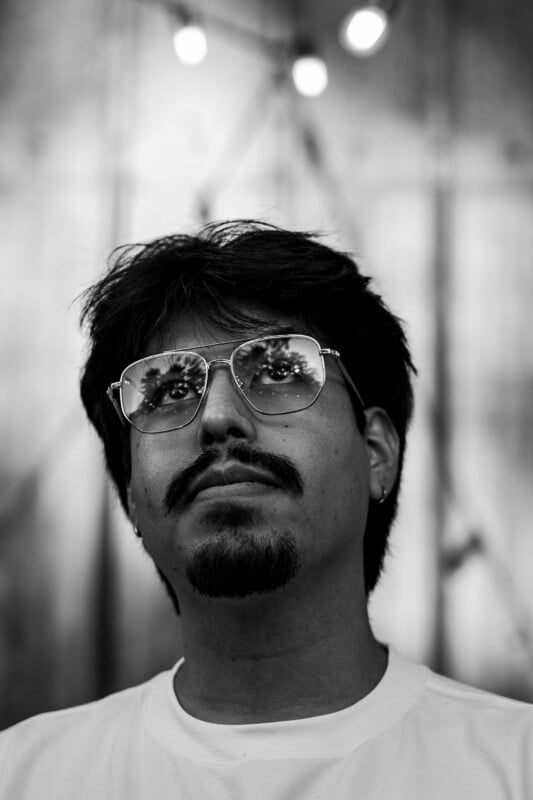 Black and white portrait of a man with medium-length dark hair, mustache, and goatee, wearing aviator glasses and a white shirt. Reflections of trees are visible in his glasses, and blurred lights glow in the background.