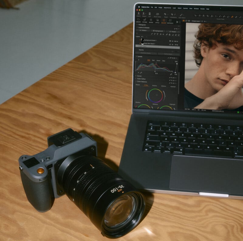 A camera with a large lens rests on a wooden table next to a laptop showing photo editing software with a portrait of a young person on the screen.