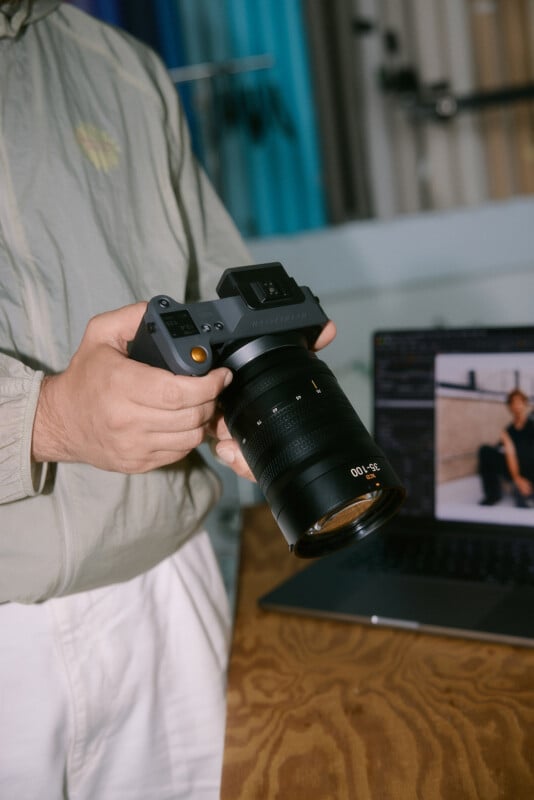 A person in a light jacket holds a Hasselblad camera with a 35-100mm lens next to a wooden table, with a laptop displaying a photo editing program in the background.