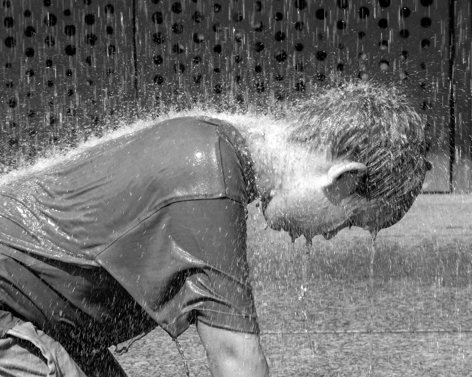 A young child in a wet t-shirt bends forward as water sprays over them, creating a curtain of droplets, with a dark perforated wall in the background.