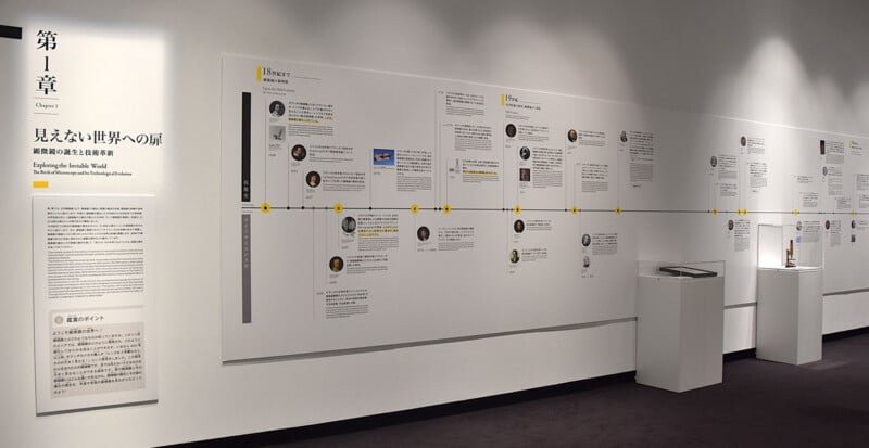 A museum exhibit wall features a long timeline titled "Exploring the Invisible World" with historical text, portraits, and two pedestals displaying microscopes.