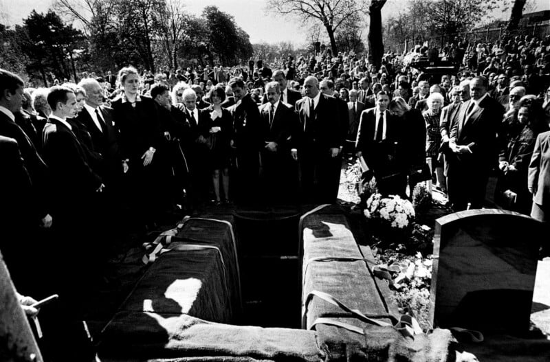 A large group of mourners, many in dark suits, stand around an open grave at a cemetery during a funeral, with a crowd gathered in the background under trees.