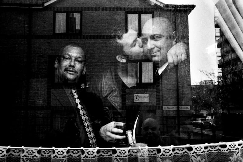Three men are seen through a window: one stands holding a drink, while two others are close together, one kissing the other's cheek. A building is reflected in the glass. A lace curtain is visible at the window’s bottom edge.