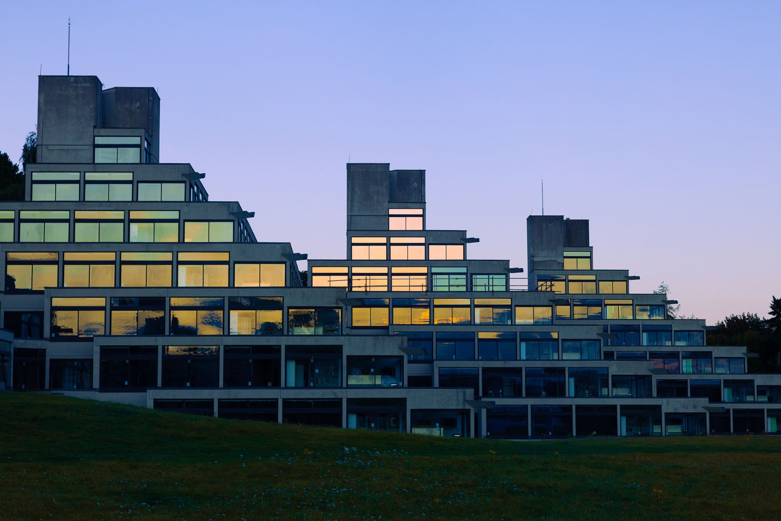 A stepped, modern concrete building with large reflective windows is shown at sunset, with a grassy foreground and a clear sky in the background.