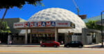 The Pacific’s Cinerama Theatre features a distinctive white geodesic dome, a retro marquee, and colorful movie posters on its façade, with palm trees and a clear blue sky in the background.