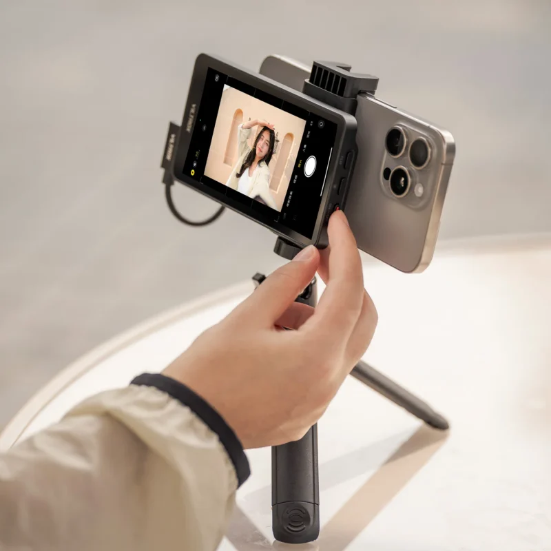 A hand holds a smartphone mounted on a small tripod, with an external monitor attached to the back of the phone displaying a live camera feed.