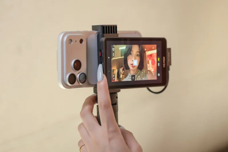 A hand holds a smartphone attached to a rear-facing external monitor display, showing a live video feed of a woman.