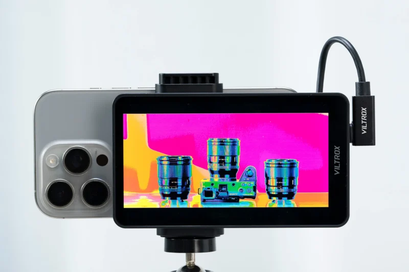 A Viltrox monitor is mounted to the back of a smartphone, displaying a high-contrast, color-inverted image of three camera lenses on a tripod.