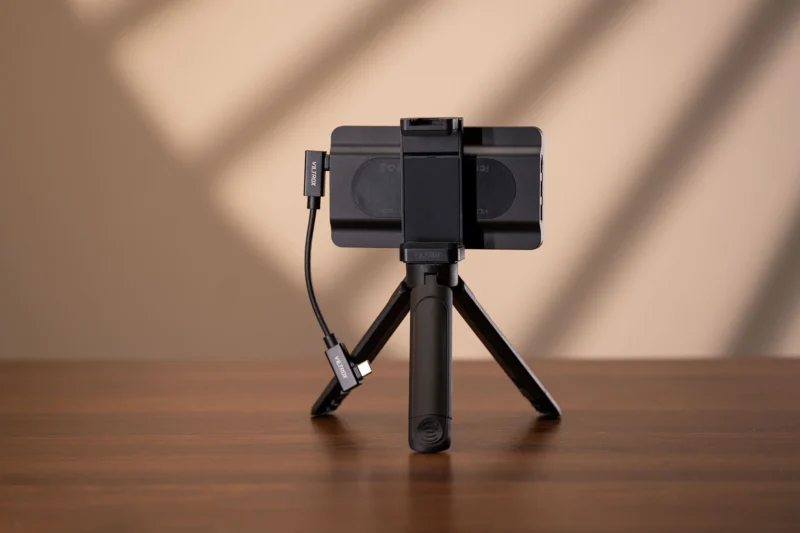 A black Viltrox smartphone mount attached to a small tripod stands on a wooden table against a beige wall with soft shadows.