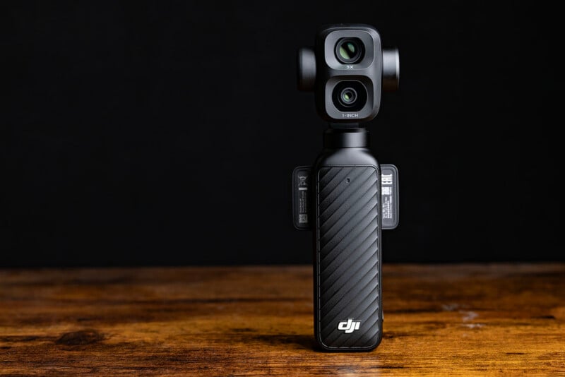 A compact DJI handheld camera with a dual-lens gimbal stands upright on a wooden surface against a black background. The device has a textured grip and visible branding on the front.