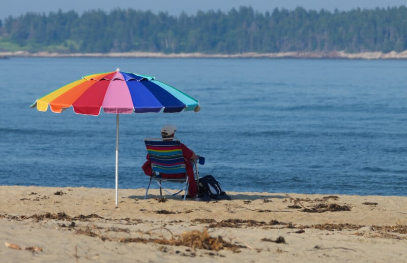 A person sits on a striped beach chair under a colorful umbrella on a sandy beach, facing the calm blue water, with a forested shoreline in the distance.
