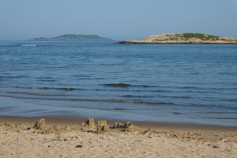 A sandy beach with small sandcastles in the foreground, gentle waves, and two green islands in the distance under a clear blue sky.