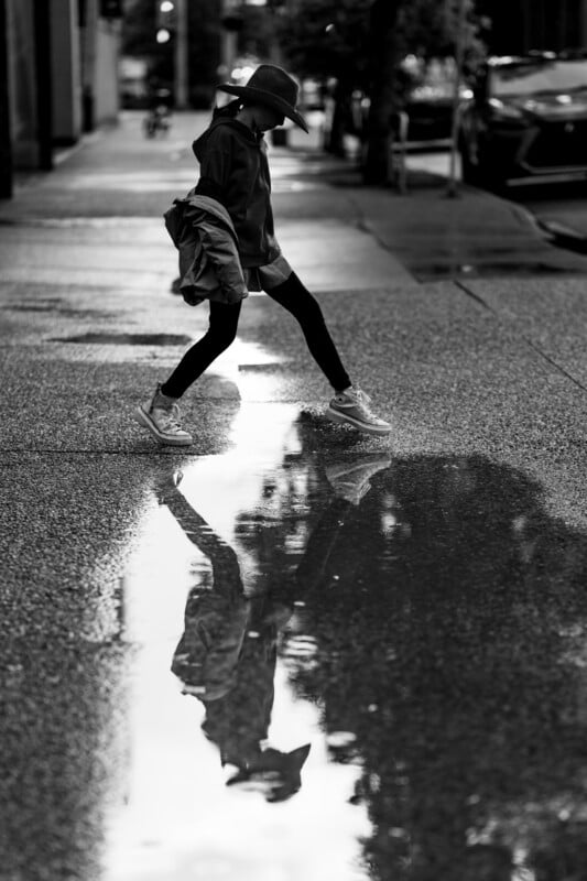 A person in a hat and coat steps over a rain puddle on a city sidewalk, their reflection visible in the water. The photo is black and white.