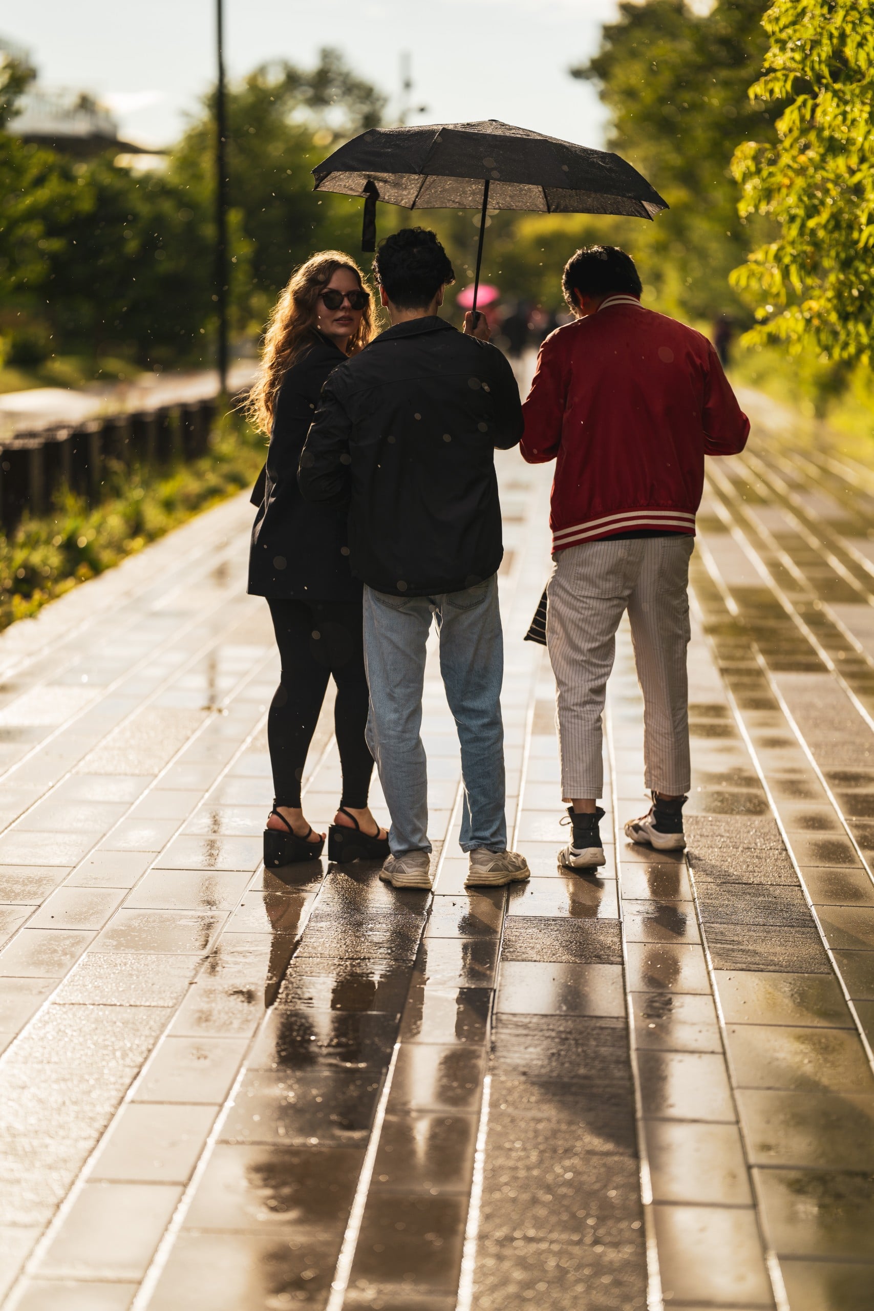 Three people stand on a wet sidewalk under an umbrella on a rainy day. One person looks back and smiles, while the others face away. Sunlight reflects off the ground.