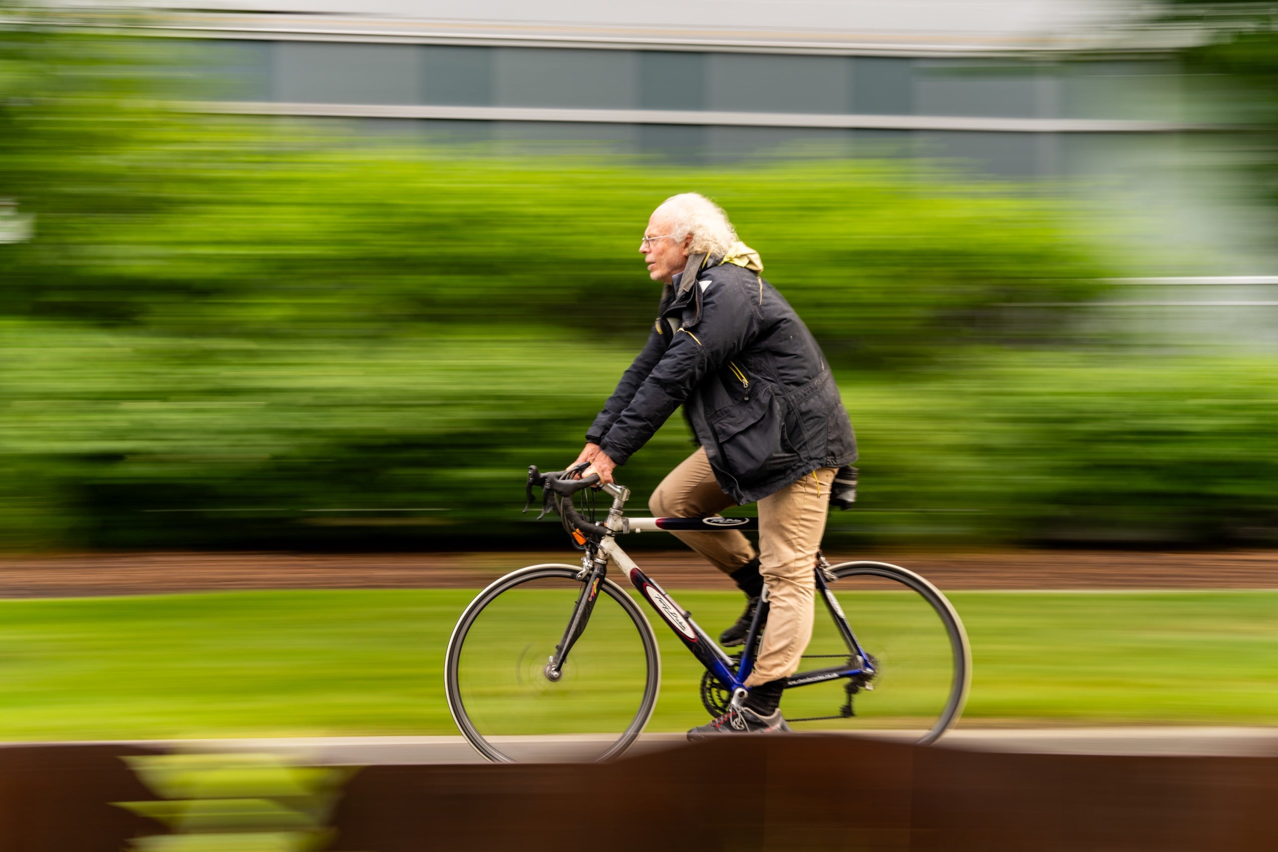 An older man with white hair rides a bicycle past green bushes, wearing a black jacket and beige pants. The background is blurred, showing motion.