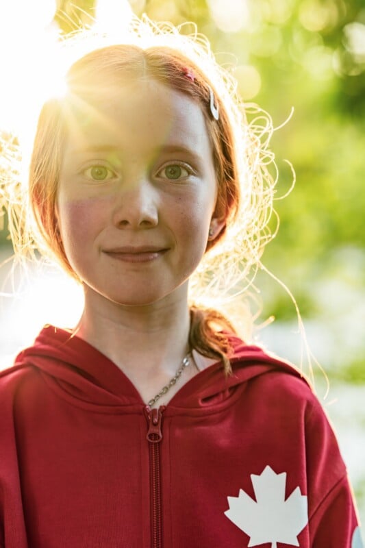 A young girl with red hair, wearing a red hoodie with a white maple leaf, stands outdoors in sunlight, smiling softly.
