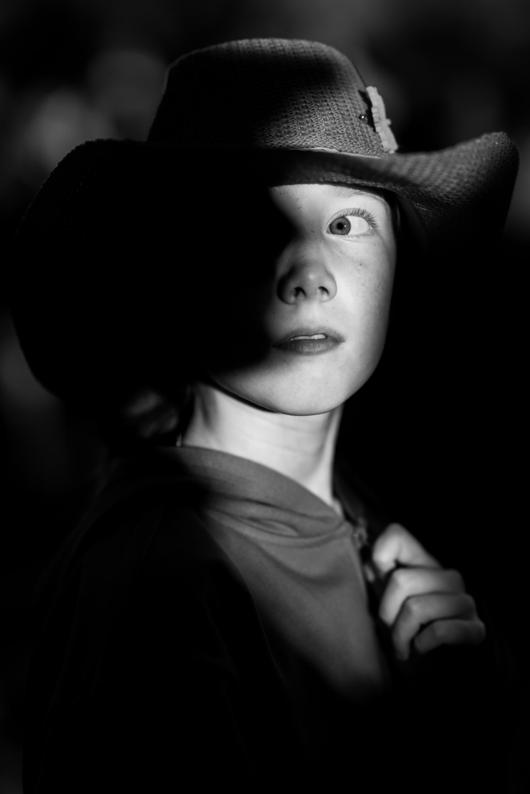A child in a wide-brimmed hat looks upward, half of their face dramatically shadowed. The lighting highlights one eye and part of their face, creating a sense of mystery. The child holds their shirt or jacket.