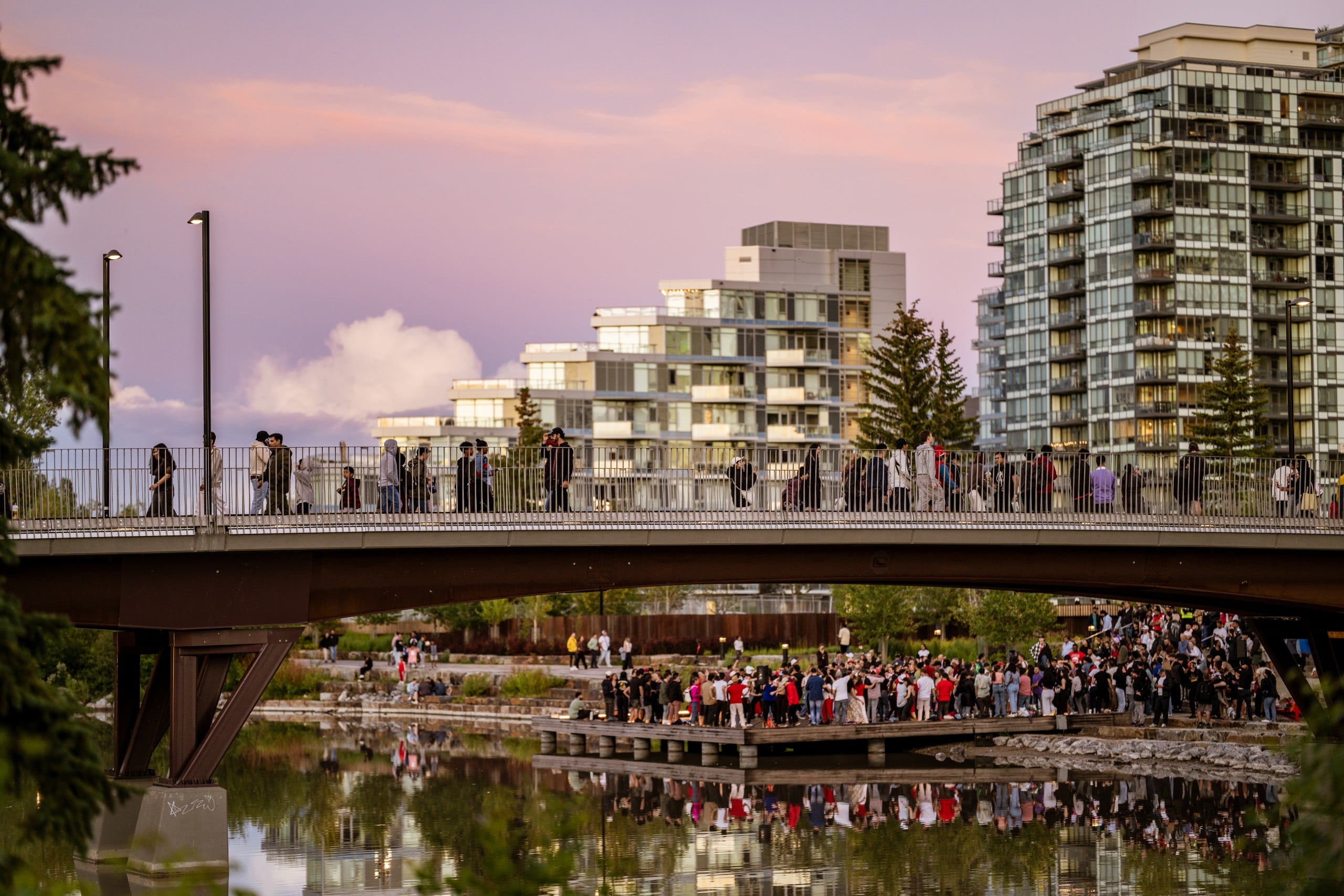 A crowd gathers on and below a bridge over calm water, with modern apartment buildings and trees in the background during sunset.