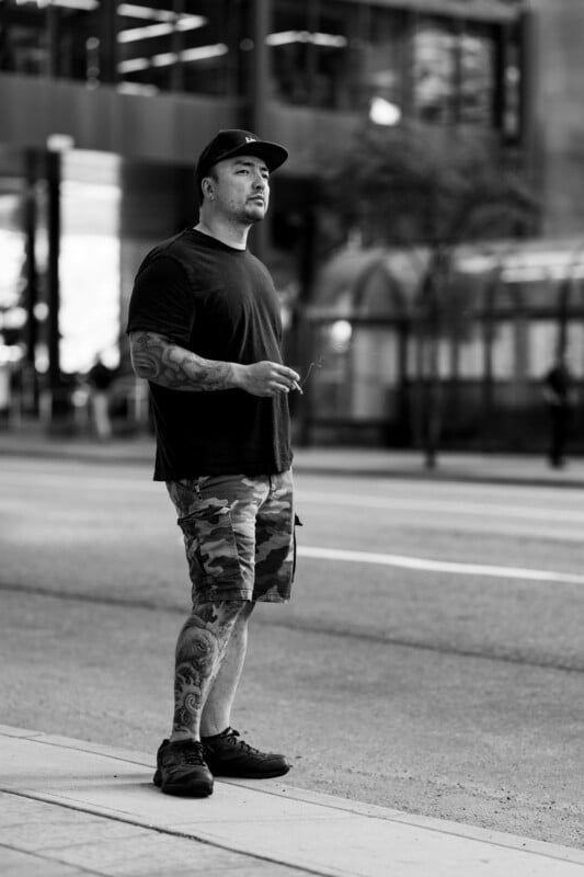 A man in a black shirt, camo shorts, and a cap stands on a city sidewalk holding a cigarette, with tattoos on his arm and leg. The background shows blurred buildings and trees. Black and white photo.