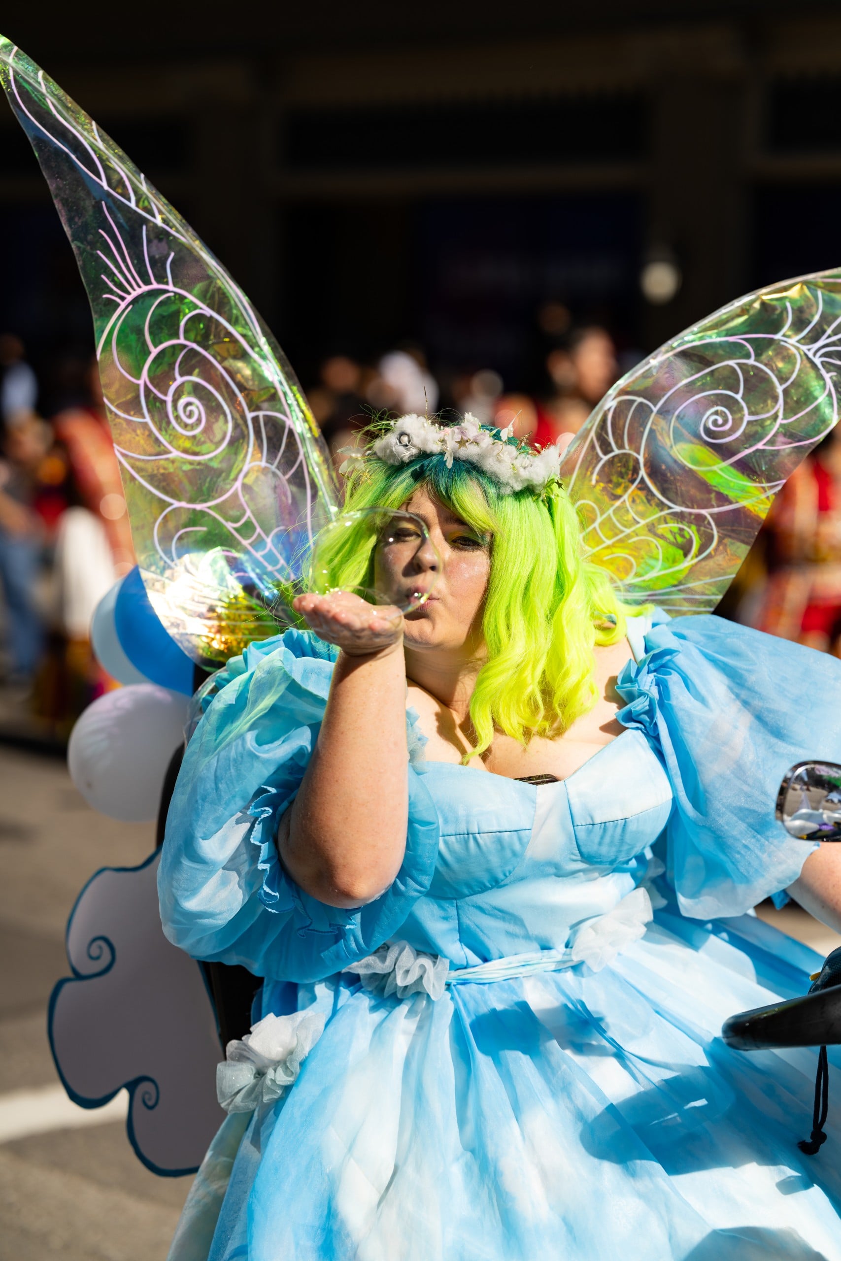 A person in a bright green wig, fairy wings, and a blue dress blows a kiss while riding in a parade. Sunlight highlights the colorful wings and festive atmosphere.