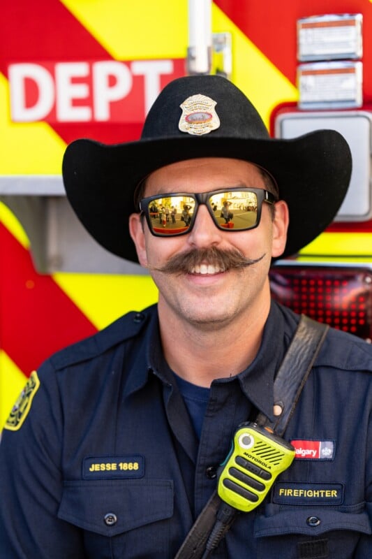A firefighter in uniform and a black hat with a large mustache smiles in front of a fire truck. He wears sunglasses, and a radio is clipped to his chest. The fire truck has bold yellow and red stripes.