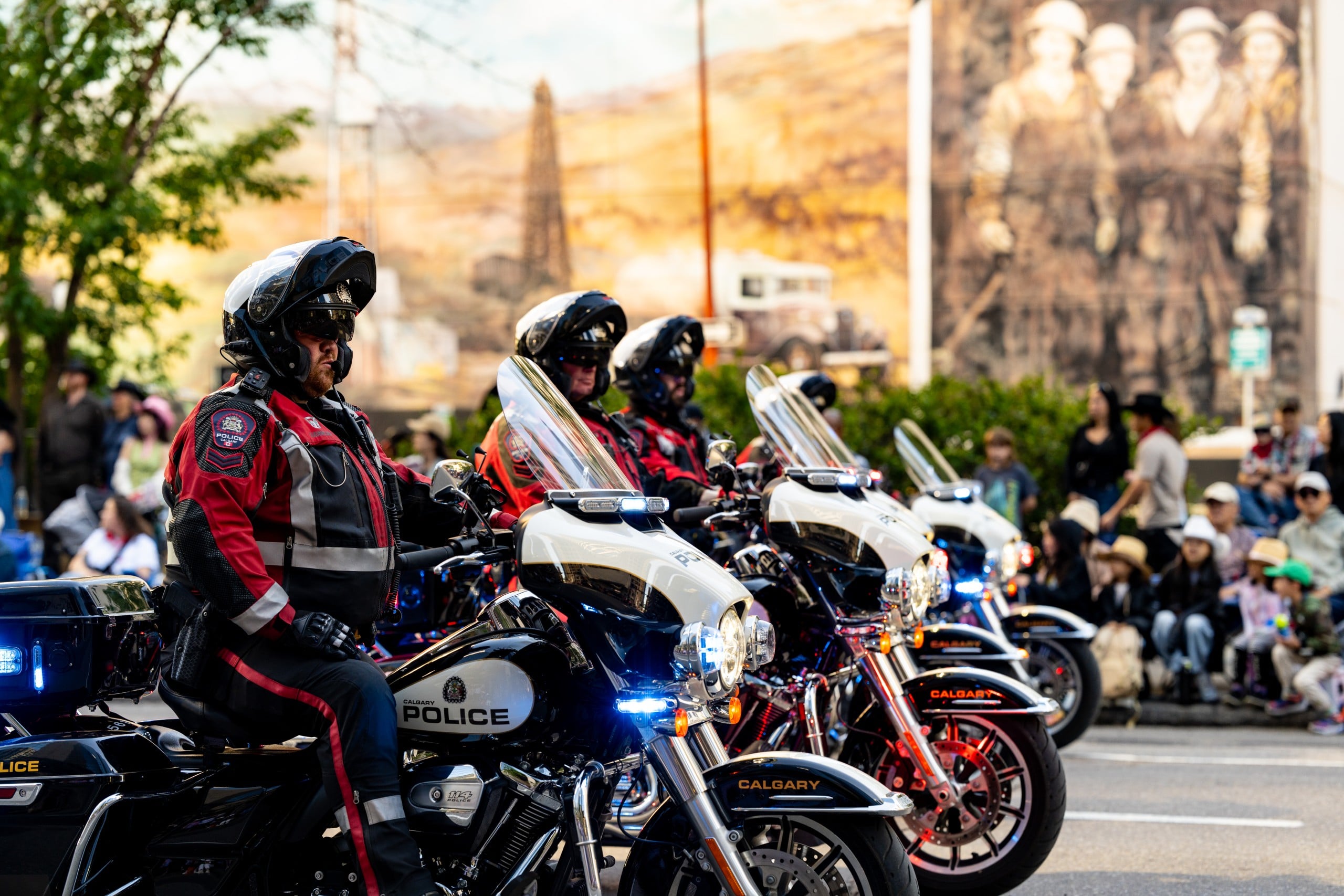 Three police officers in uniform ride motorcycles during a parade, with a crowd watching and a mural of historical figures in the background.