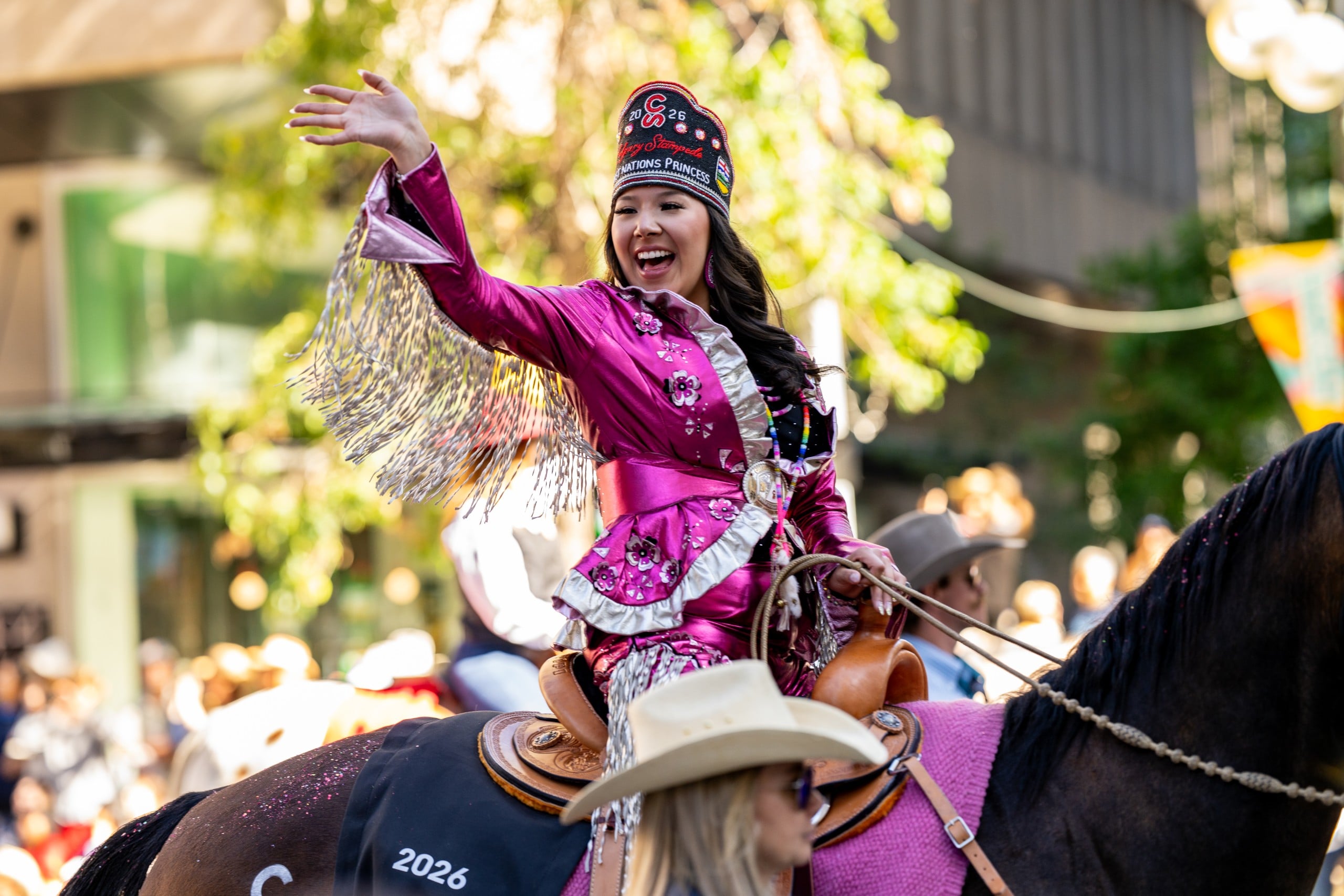 A woman in a shiny pink rodeo outfit and crown waves while riding a horse in a parade. People in cowboy hats watch in the background. The scene is lively and festive.