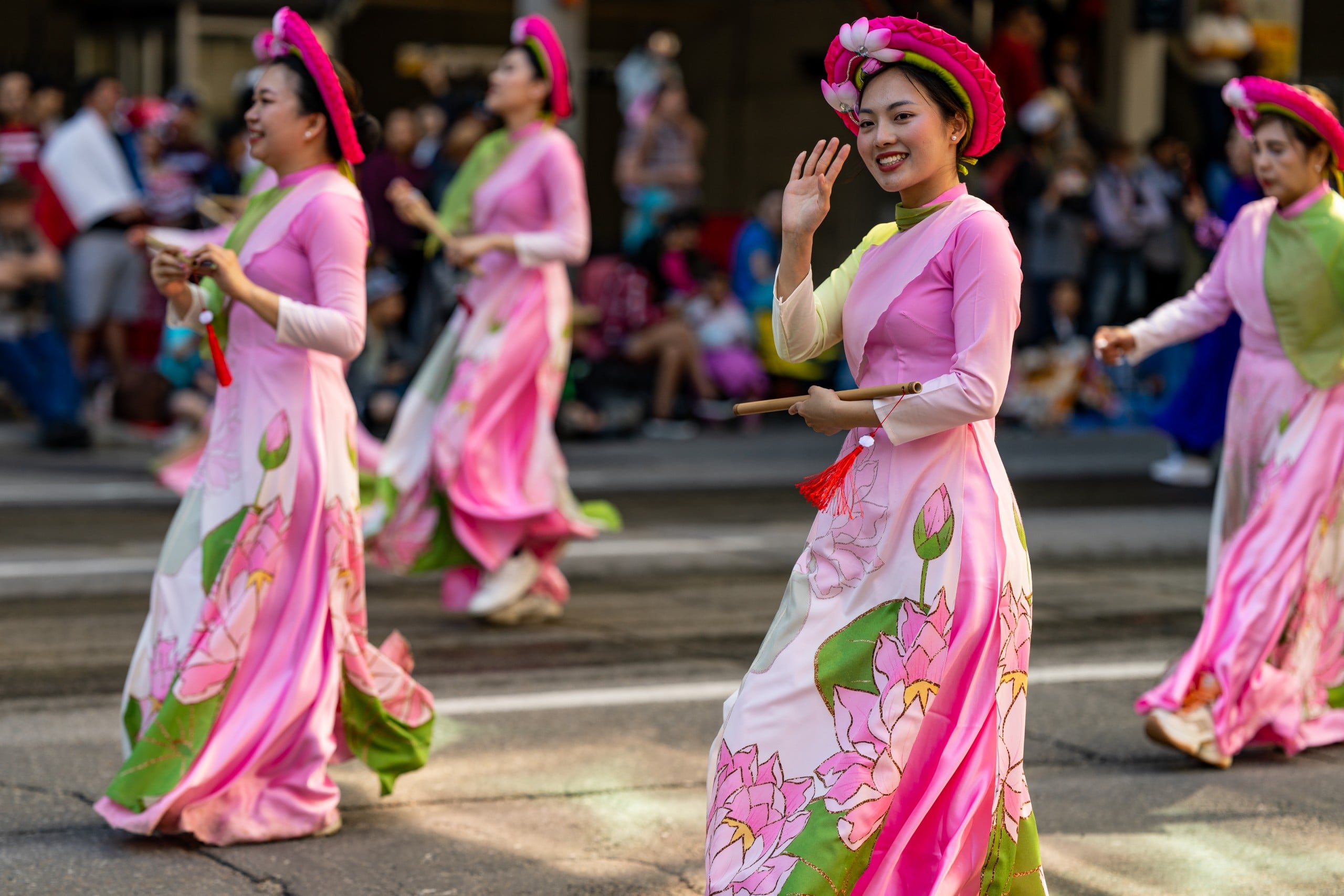 Women in pink traditional dresses with flower patterns walk in a parade. One woman in front smiles and waves at the camera. Spectators watch from the background.