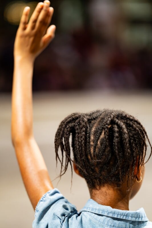 A child with braided hair, seen from behind, raises their right hand while wearing a blue shirt. The background is blurred.