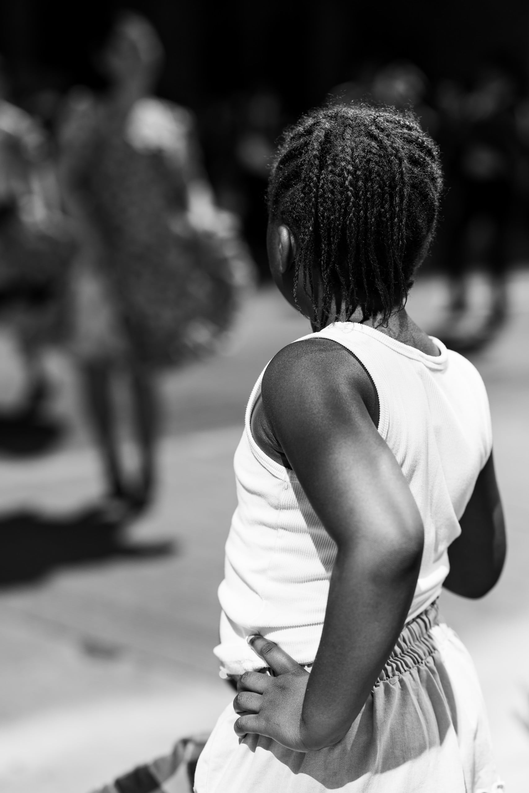 A young girl with braided hair stands with her hands on her hips, facing away from the camera. She wears a sleeveless top and looks toward a blurry crowd in the background. The image is in black and white.