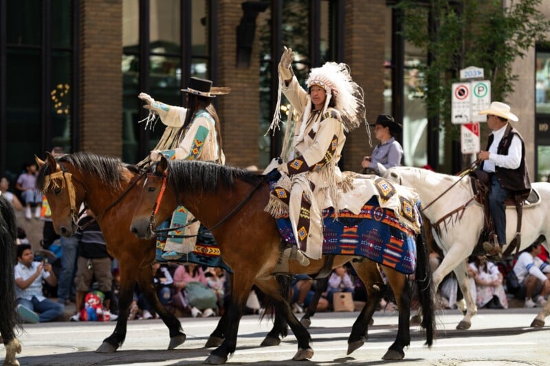 Three people in traditional and western clothing ride decorated horses in a parade. The crowd watches from the sidewalk, and city buildings and signs are visible in the background.