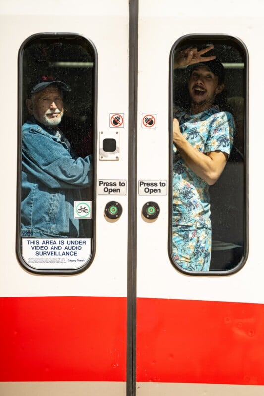 Two men stand inside a train behind closed doors. One is older, wearing a cap and jacket; the other, younger, wears a colorful shirt and flashes a peace sign. Surveillance and no smoking signs are visible on the doors.