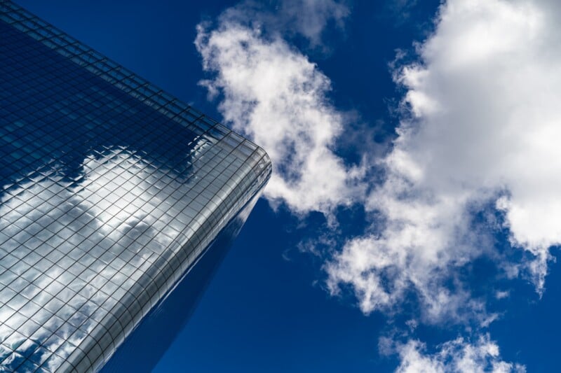 A modern glass skyscraper with clouds reflected on its surface stands against a bright blue sky with white clouds.