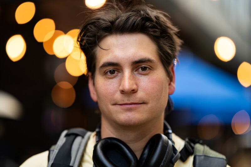 A young man with tousled brown hair wears headphones around his neck and a backpack, standing outdoors with warm blurred lights in the background.