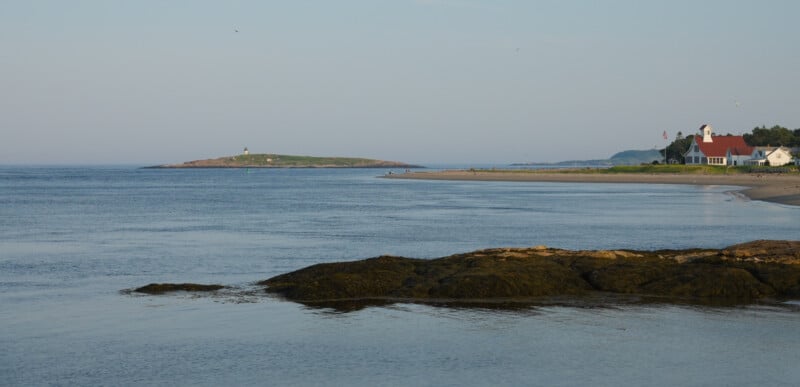 A calm coastal scene with gentle waves, rocky shoreline in the foreground, a sandy beach, a white house with a red roof, and a small island with a lighthouse in the distance under a clear sky.