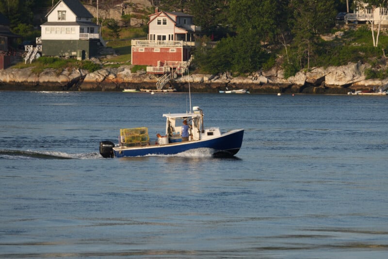 A small blue fishing boat with lobster traps on board moves across calm water near a rocky shoreline with houses and trees in the background.