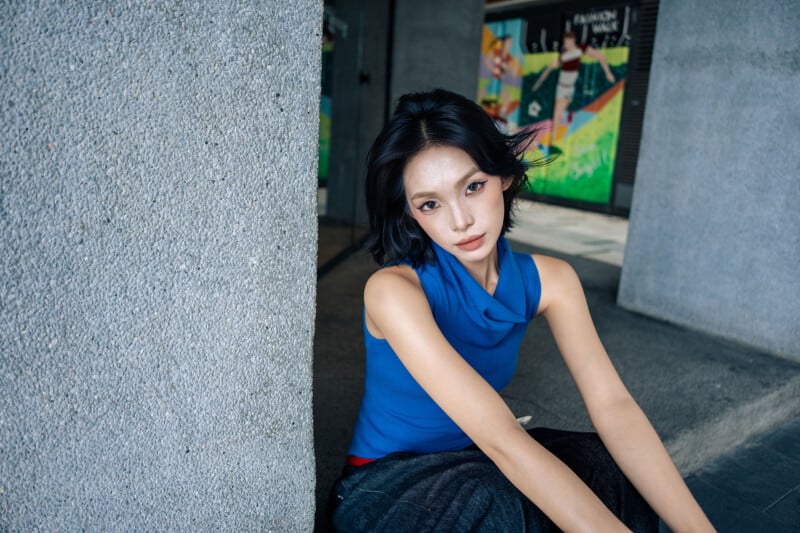 A woman with short dark hair and a blue sleeveless top sits on the ground, leaning against a concrete wall, with colorful posters visible in the background.
