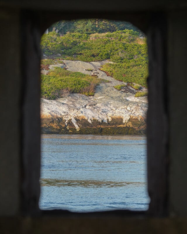 View of rocky shoreline and green foliage across calm water, framed by a rectangular stone window or opening.