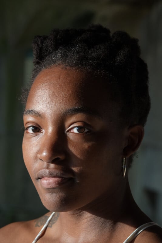 A person with short, natural hair and hoop earrings gazes into the camera, illuminated by soft sunlight, with a neutral expression and a blurred background.