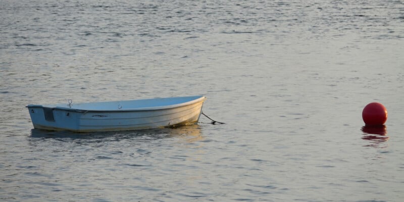 A small white rowboat floats on calm water near a bright red buoy, with gentle ripples reflecting the soft light.