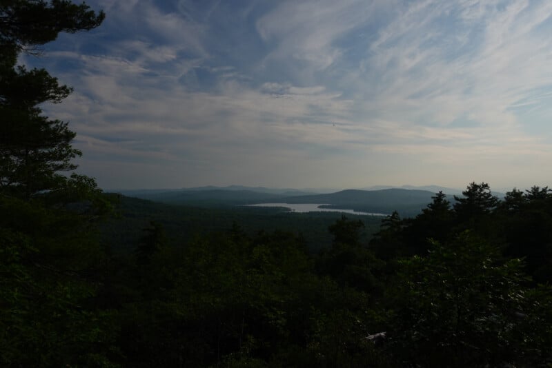 A scenic view of a forested landscape with a lake in the distance, surrounded by rolling hills under a sky with wispy clouds. Trees frame the foreground.