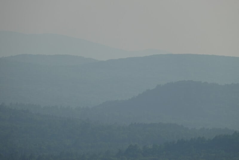 Fog covers a landscape of rolling, tree-covered hills with layers of distant blue-green mountains fading into the hazy sky.