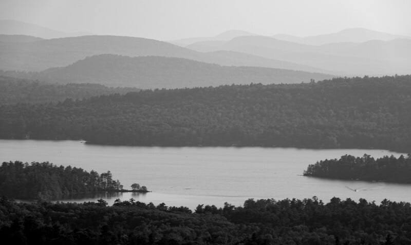 Black and white photo of a calm lake surrounded by dense forests, with layers of tree-covered hills and mountains in the background under a hazy sky.