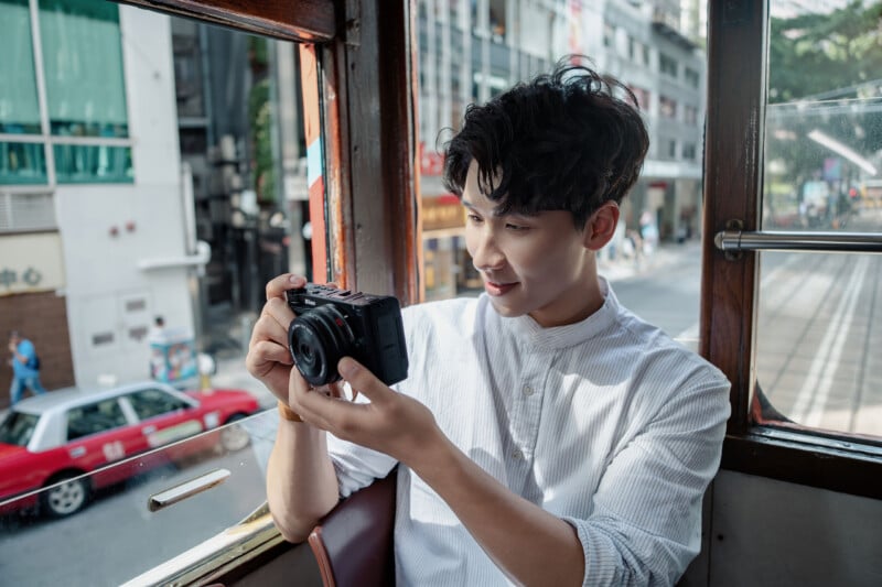 A man in a white shirt sits by a window on public transport, smiling as he looks at a digital camera. A city street with buildings and a red taxi is visible outside the window.