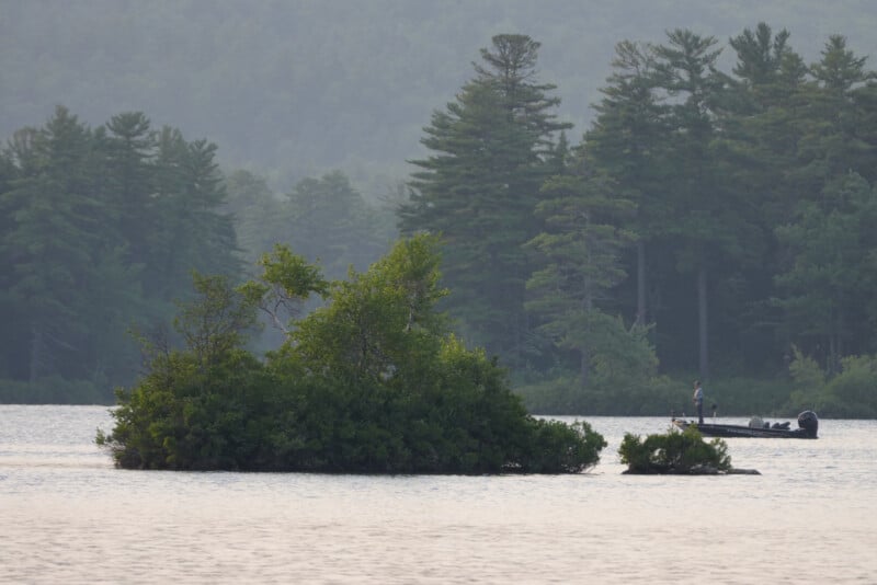 A small, green island in a calm lake with a person standing on a boat in the background, surrounded by tall pine trees and forested hills.