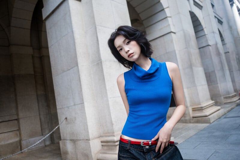 A person with short black hair wears a blue sleeveless top, jeans, and a red belt, standing in front of stone arches on a city sidewalk.