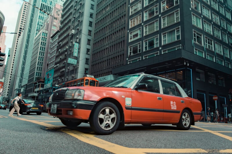 A red taxi drives through an urban street intersection surrounded by tall buildings, with a few pedestrians crossing and city traffic in the background.