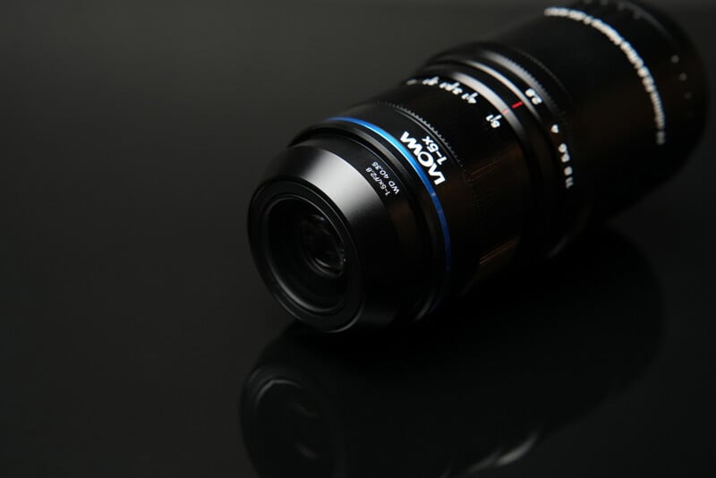 A black Laowa 1-5x macro lens sits on a reflective black surface, showing its magnification markings and aperture ring.