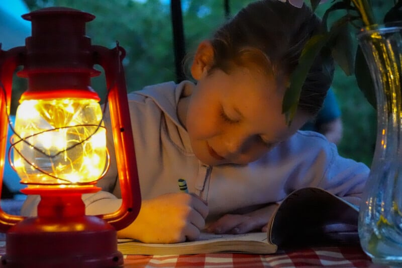A young girl writes in a notebook by the light of a glowing red lantern on a checkered table, with a glass vase and greenery nearby, creating a cozy evening scene.